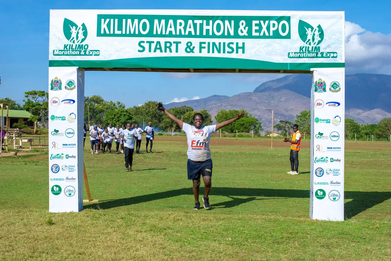 A runner throwing his arms wide under the Kilimo Marathon arch, the Uluguru mountains behind